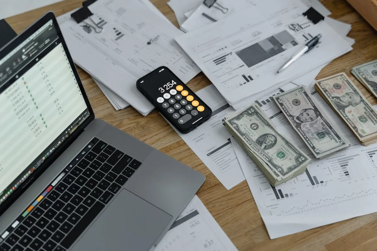 Laptop with spreadsheet, calculator app, and cash on a desk with financial reports